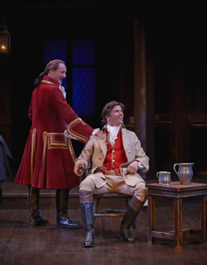 Archer in long red coat with gold embroidery and riding boots, his hair in a pony tail, leans on  the chair in which sits Aimwell, in tan coat and britches, red vest, and fluffy white shirt, black riding boots with brown tops, and long hair. Next to him is a table with pewter ewer and tankards